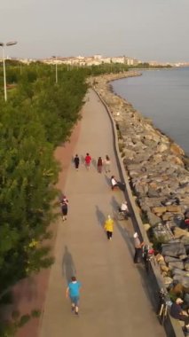 Sahil boyunca bir sahil şehrinin üzerinde uçmak, yukarıdan şehrin manzarası, deniz kenarındaki koşu bandında yürüyen insanlar