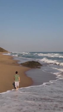 Boş kumsalda, dalgalı denizde, boş koylarla, yeşil tepelerin arkasındaki denizde yürüyen bir adamın insansız hava aracı görüntüsü.