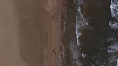 Denizin sahile vuruşunu ve kumsalda yürüyen iki kişinin uzun sahildeki dalgaların görüntüsünü.