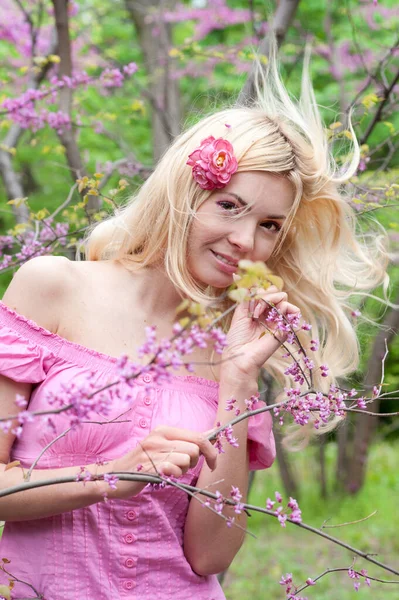 幸せなブロンドふわふわ髪の女性ポーズで春の開花公園 かわいいピンクのドレスを着て — ストック写真