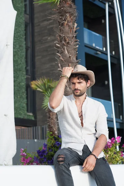 Young serious handsome man outdoor portrait, man dressed in hat and white shirt posing in tropical city