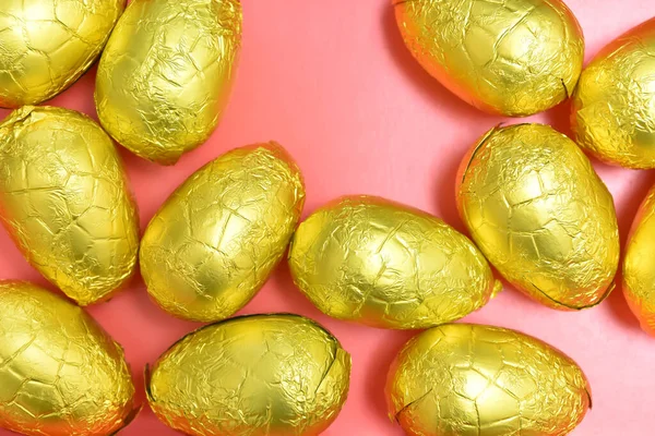 Pile or group of gold, or yellow foil wrapped chocolate easter eggs, against a pink, peach orange background.