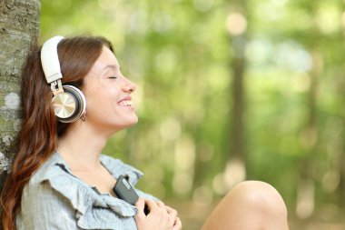Parkta ya da ormanda kulaklıkla müzik dinleyen mutlu bir kadının yan görüntüsü.
