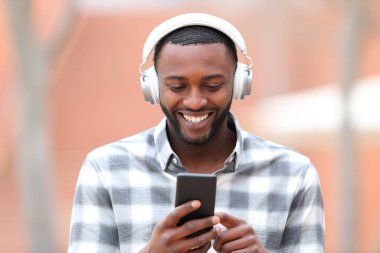 Siyahi bir adamın sokakta kulaklık ve cep telefonuyla müzik dinlemesini ön planda görmek.