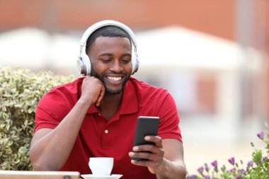 Happy black man in red listening to music wearing headphones checking phone in a coffee shop