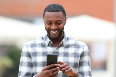 Front view portrait of a happy black man walking using smart phone in the street