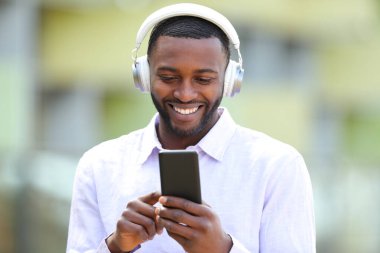 Front view portrait of a happy black man wearing wireless headphones listening to music online on phone