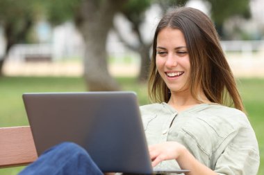 Happy teen using laptop sitting on a bench in a park