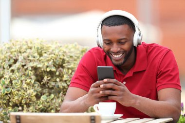 Happy black man and red shirt listening to music with headphones and phone sitting in a bar terrace