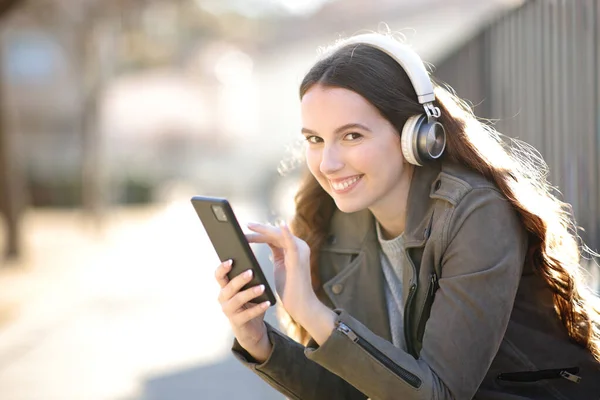 幸せな女性はヘッドフォンを身に着けているあなたを見て通りにスマートフォンでオーディオを聞く — ストック写真