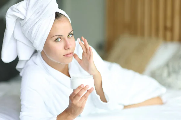 用润肤霜洗脸的女人看着相机洗澡后躺在床上 — 图库照片