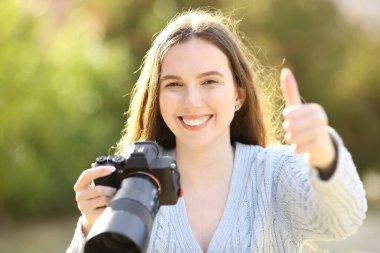 Parkta başparmak hareketi yapan aynasız fotoğraf makinesi tutan mutlu bir fotoğrafçının ön manzara portresi.