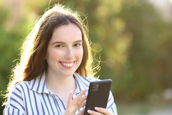 Szczęśliwa Kobieta Trzyma Telefon Komórkowy Patrzy Kamerę Stojącą Parku — Zdjęcie stockowe