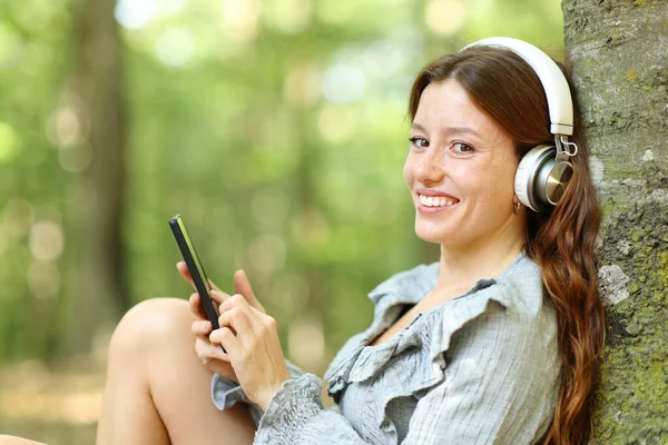 ヘッドフォンと電話で音楽を聴いている幸せな女性は森の中であなたを見て — ストック写真