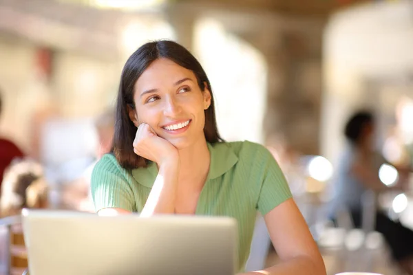 Happy woman with laptop thinking in a restaurant terrace looking at side