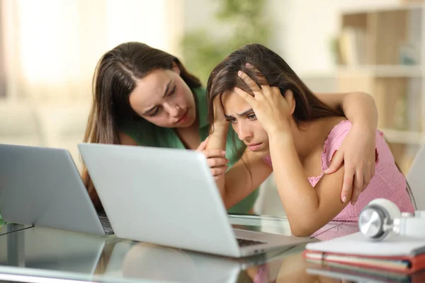 Sad student being comforted by another one checking laptop at home