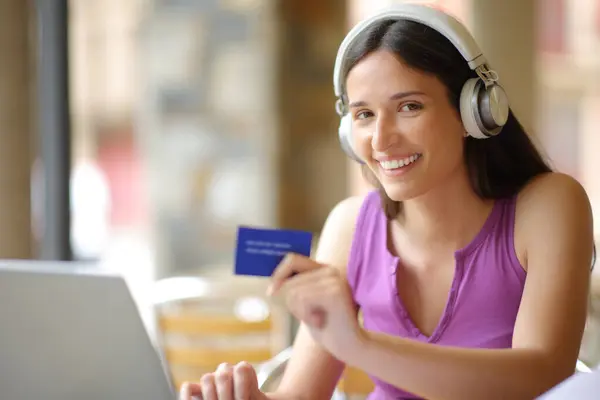 Happy buyer wearing headphone with laptop and credit card buying media online looking at you in a restaurant terrace