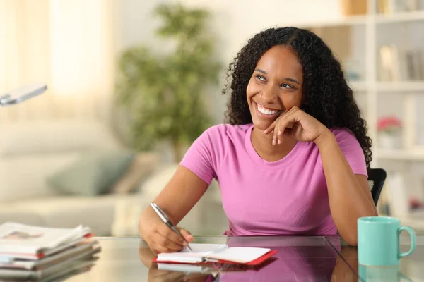 Happy black woman writing in agenda dreaming looking at side at home