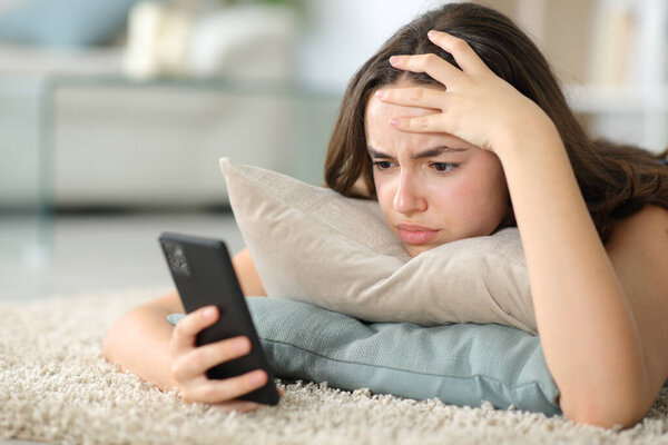 Worried woman checking phone lying on the floor at home