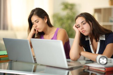 Evde dizüstü bilgisayarlarıyla online içeriği bekleyen iki sıkılmış öğrenci.