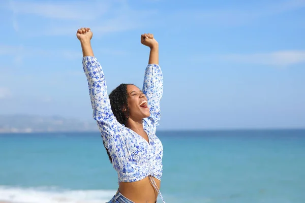 Excited black woman raising arms celebrating on the beach a sunny summer day