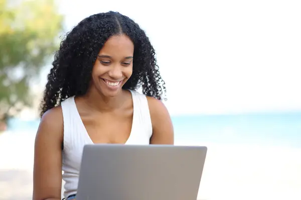 快乐的黑人女人坐在沙滩上检查笔记本电脑 图库图片