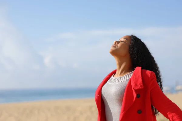 在阳光明媚的日子 穿着红色夹克的黑人妇女在海滩上呼吸新鲜空气 免版税图库照片