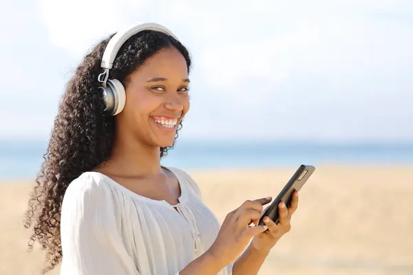 Happy black woman listening audio holding phone on the beach looking at camera a summer sunny day