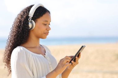 Güneşli bir günde kumsalda yürüyen akıllı telefon kullanan siyahi kadın.