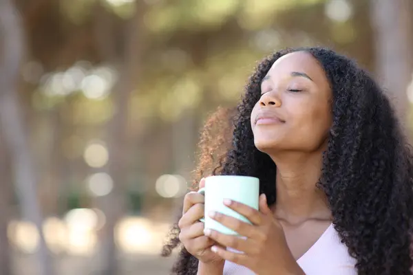 站在公园里的黑人妇女从杯子里闻到咖啡的味道 免版税图库照片