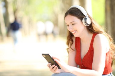 Mutlu kadın kulaklık takıyor ve cep telefonunu kontrol ediyor. Parkta müzik dinliyor.