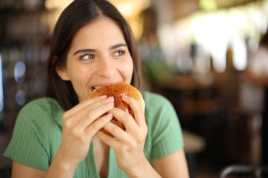 Bir restoranın içinde hamburger yiyen mutlu bir kadın.