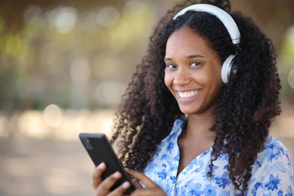 Kulaklık takan mutlu zenci kadın parkta telefon dinlerken sana bakıyor.