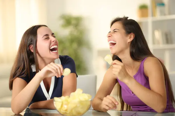 Two happy friends laughing hilariously eating potato chips at home