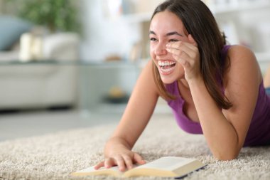 Happy woman laughing hilariously reading a comedy paper book lying on the floor at home clipart