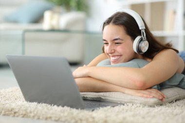 Happy woman watching media content on laptop lying on a carpet at home clipart