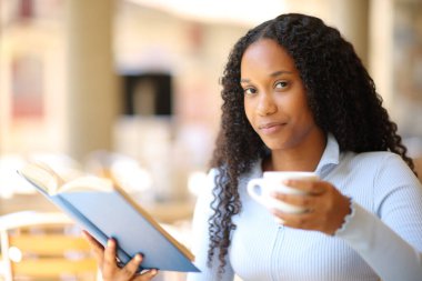Kâğıt kitap ve kahve fincanı tutan mutlu siyah kadın restoranın terasında sana bakıyor.