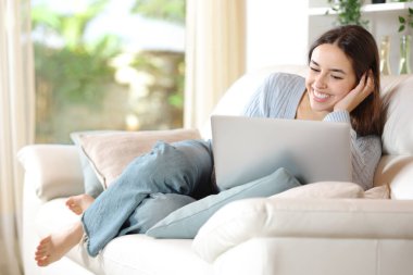 Full body portrait of a happy woman using laptop sitting on a couch at home clipart