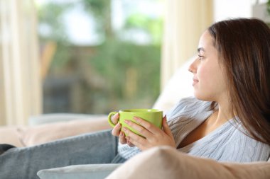 Elinde kahve fincanı olan rahat bir kadın evdeki pencereden bahçeyi inceliyor.