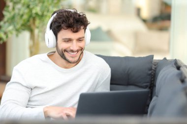 Kulaklık takan mutlu bir adam dizüstü bilgisayarda medyayı izliyor terasta bir kanepede oturuyor.