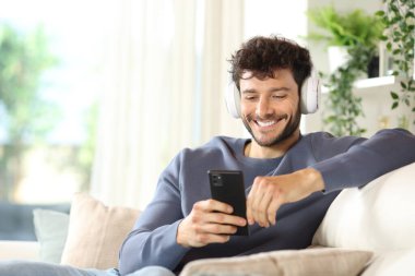 Kulaklık takıp müzik dinleyen mutlu bir adam telefonuyla evdeki bir kanepede oturuyor.