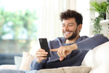 Evin içindeki bir koltukta oturan akıllı telefonu kullanan mutlu bir adam.
