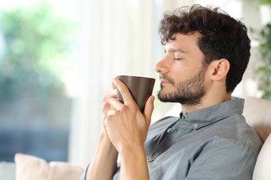 Profile of a relaxed man smelling coffee sitting in a house interior clipart