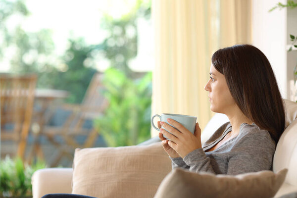 Serious woman holding coffee cup looking at garden through a window at home