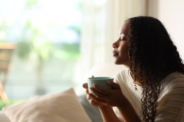 Evde kahve içen ve gözleri kapalı rahatlayan siyah bir kadının yan görüntüsü.