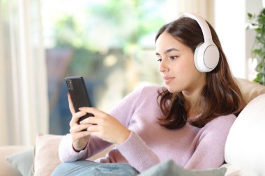 Kanepede oturan, kulaklık takan, evde telefonla medyayı izleyen bir kadın.
