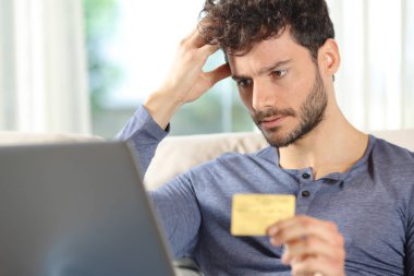 Kredi kartı ve dizüstü bilgisayarla internetten alışveriş yapan endişeli bir adam evde şikayet ediyor.