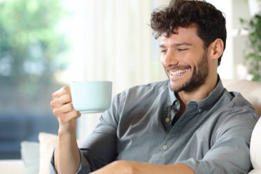 Evdeki kanepede oturan mutlu adam kahve fincanına bakıyor.