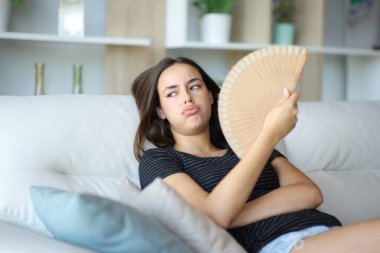 Overwhelmed woman using fan in a hot summer day sitting on a couch at home clipart