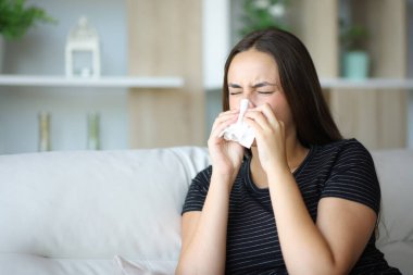 Evde kanepede oturan, mendil kullanarak hapşıran hasta bir kadın.